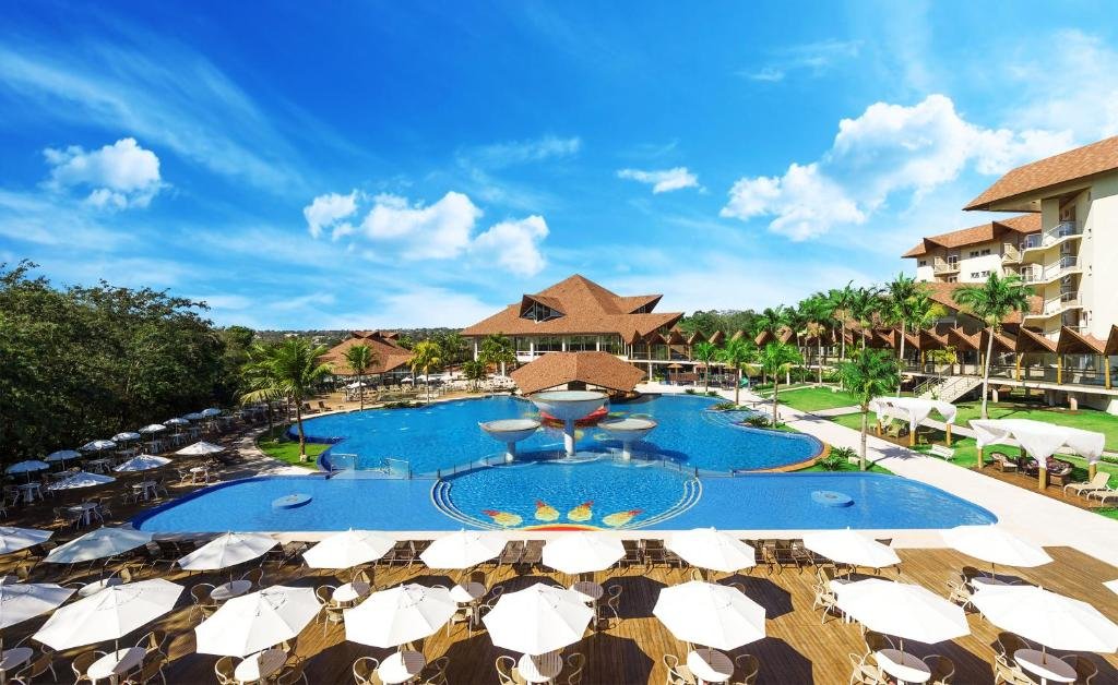 Parte do Recanto Cataratas que mostra uma grande piscina ao ar livre com um quiosque no meio e várias mesas com cadeiras e guarda-sóis em volta. Ao fundo tem algumas árvores e parte da construção do resort. Imagem ilustrando post Resorts em Foz do Iguaçu.