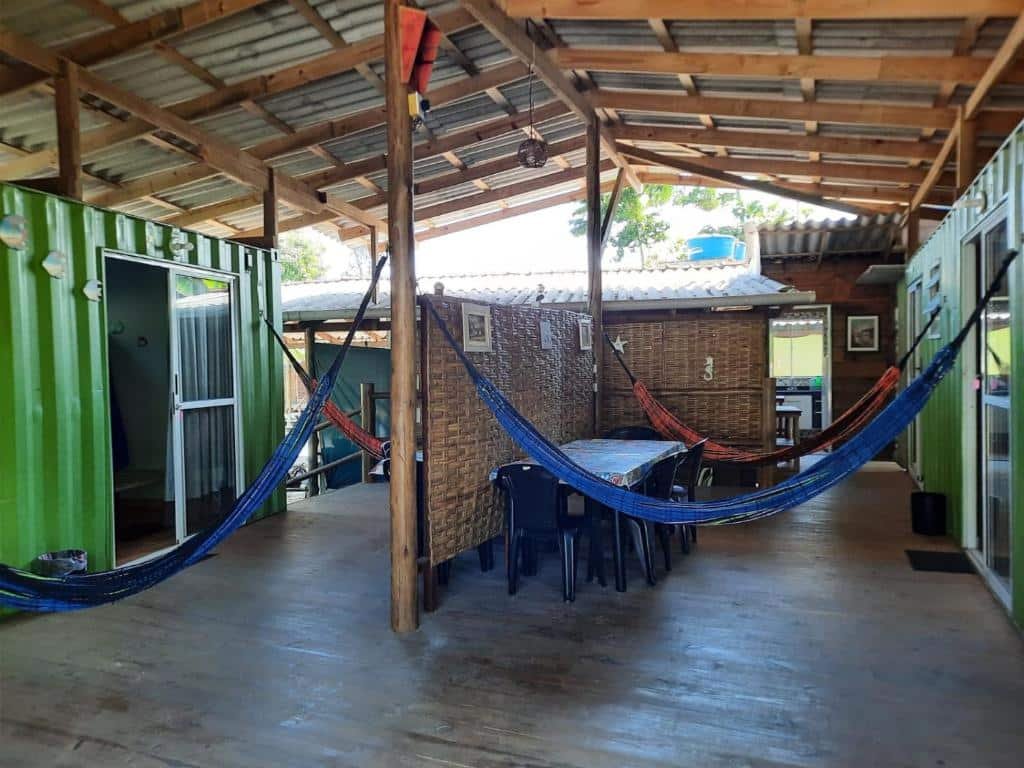 Área externa do Recanto do Estaleiro. Duas redes de cada lado, uma parede no meio com uma mesa e cadeiras, de cada lado portas dos quartos.