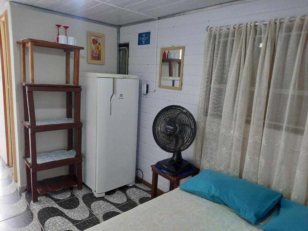 Quarto do Recanto do Estaleiro. Do lado direito uma cama de casal, em cima uma janela, do lado um banco com um ventilador. Do lado esquerdo uma estante e uma geladeira com espelho. Foto para ilustrar post sobre airbnb em Ubatumirim.