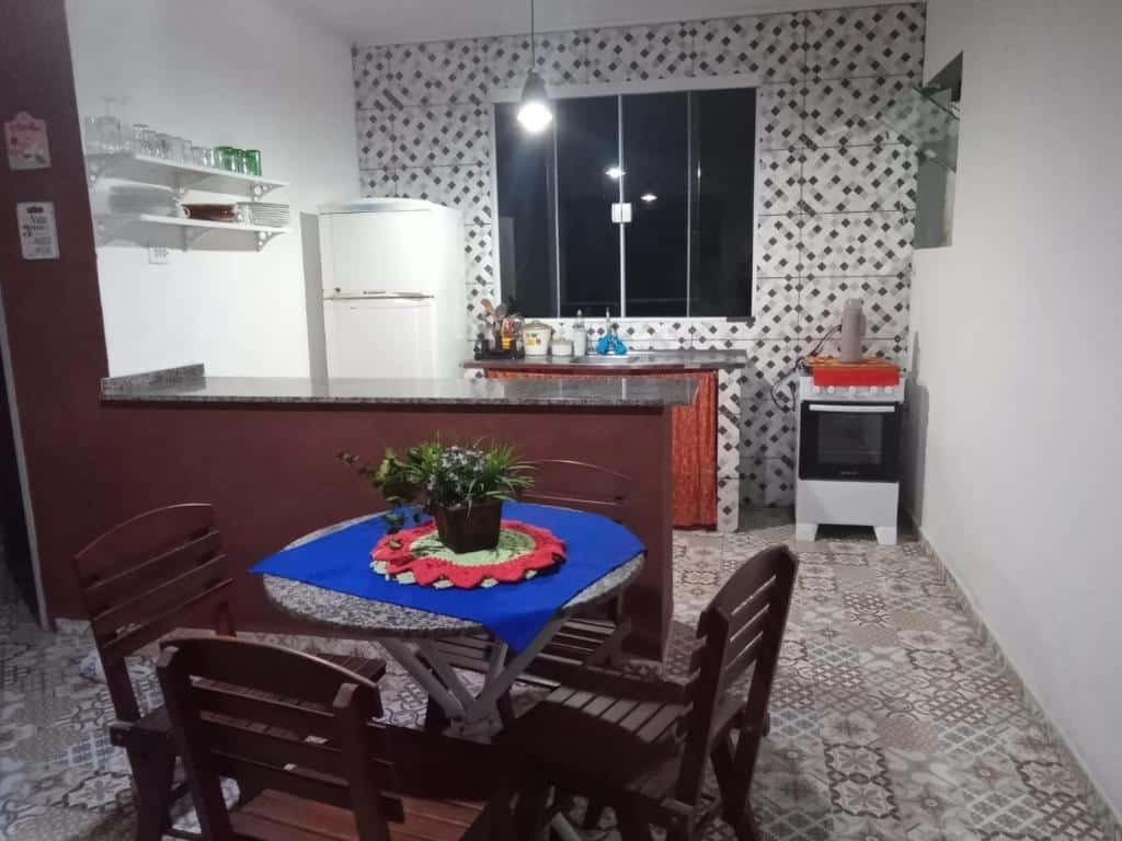 Cozinha do Recanto dos Guedes. Uma mesa com quatro cadeiras na frente, no meio um balcão. No fundo, do lado direito um fogão, no meio a pia e do lado esquerdo a geladeira. Em cima da pia uma janela.
