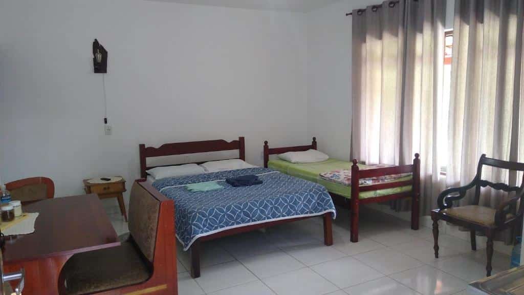 Foto do quarto em Recanto Só Alegria. É espaçoso possui uma mesa com cadeira à esquerda, uma cama de casal ao centro e outra de solteiro ao seu lado, na direita. A janela está ao lado da cama de solteiro.
Ilustra o post sobre airbnb em Serra Negra.