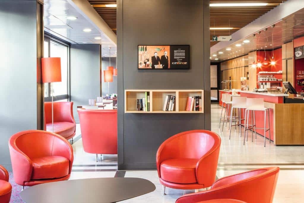 recepção do Ibis Roma Fiera com mesa cinza e cadeiras vermelhas com uma parede no meio da sala com tv e algumas revistas embaixo, do outro lado da sala, a bancada da recepção, também com detalhes vermelhos