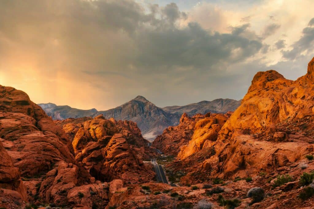 Vista das formações rochosas no Red Rock Canyon. Elas têm um tom avermelhado e estão em volta de uma estrada de asfalto (Scenic Drive). Ao fundo está o horizonte com montanhas e o céu. Esse é mais um dos pontos turísticos em Las Vegas.