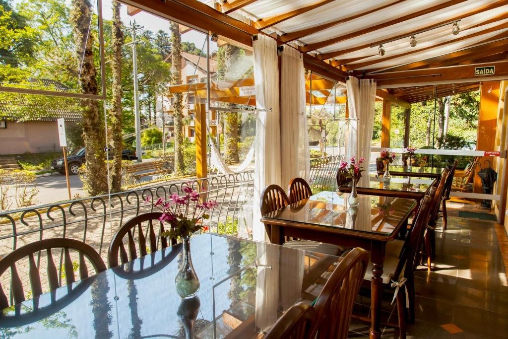 refeitório da Pousada Águia Branca em Gramado com vista para o jardim e para rua. As paredes são envidraçadas e amplas, e do lado de dentro do refeitório há várias mesas retangulares com quatro cadeiras de madeira em cada