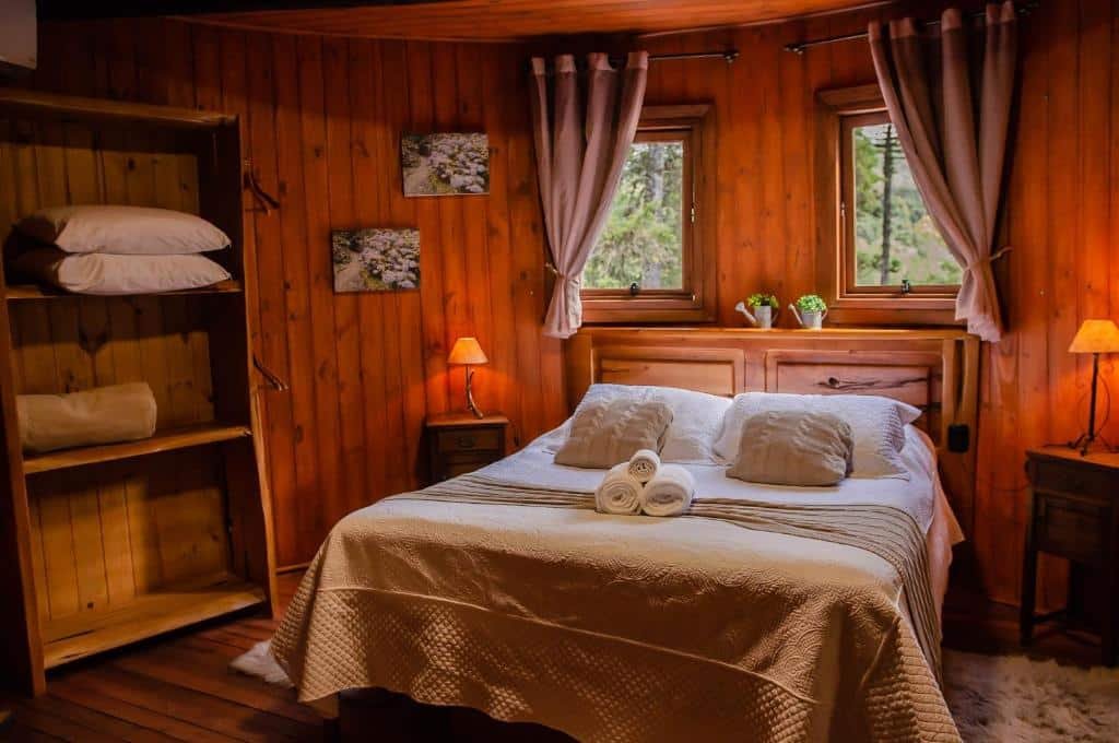 Quarto do Refúgio do Caracol Pipas e Chalés. As paredes, o chão e o teto são todos de madeira, criando uma atmosfera quente e rústica. No centro do quarto, há uma cama de casal com roupa de cama branca e duas toalhas enroladas aos pés da cama.

Ao lado da cama, há mesas de cabeceira com abajures combinando. Acima da cabeceira, há uma janela que permite a entrada de luz natural, ladeada por cortinas. Em frente à cama, há uma estante aberta com cobertores ou toalhas dobradas e itens decorativos.

Nas paredes, há quadros ou obras de arte que complementam a decoração. No chão, ao lado da cama, há um pequeno tapete, adicionando um toque extra de conforto ao ambiente.