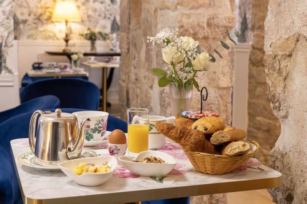 A imagem mostra uma mesa de café da manhã elegante em um ambiente acolhedor no Relais Hôtel du Vieux Paris. Sobre a mesa, há uma cesta de pães com croissants e baguetes, uma xícara de chá com pires, uma chaleira de prata, um copo de suco de laranja, um ovo cozido e tigelas com frutas frescas e cereais. Ao fundo, um vaso com flores brancas adiciona um toque de delicadeza. A parede de pedra e as cadeiras de veludo azul realçam a decoração charmosa e aconchegante do local.
