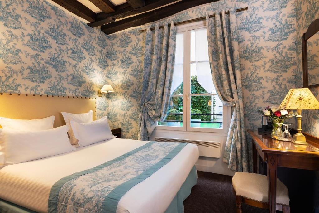 Quarto do Relais Hôtel du Vieux Paris, um dos hotéis com café da manhã em Paris. Uma cama de casal está encostada na parede esquerda do local com mesinhas de cabeceira e abajures dos dois lados. Logo em frente fica uma mesa com banquinho, abajur, vaso de flores e cafeteira. Na lateral esquerda há uma janela com cortinas.
