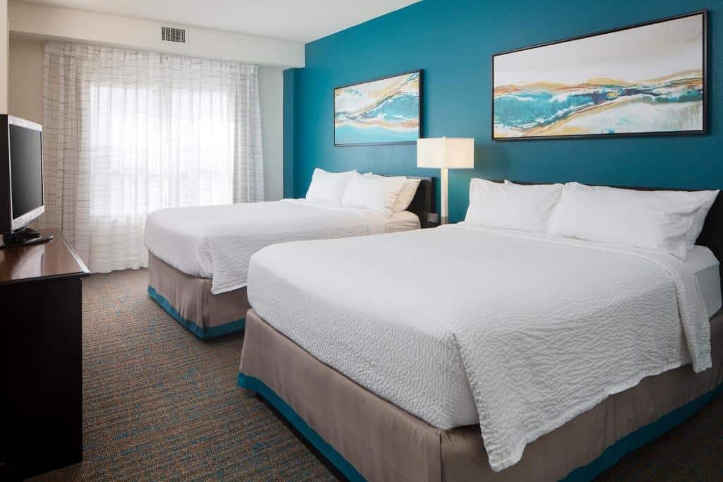 quarto do Residence Inn by Marriott Orlando com duas camas de casal, cada uma com um quadro acima e uma luminária grande de chão entre as duas e uma mesinha, na frente, há um balcão com tv em cima, janela e cortinas claras