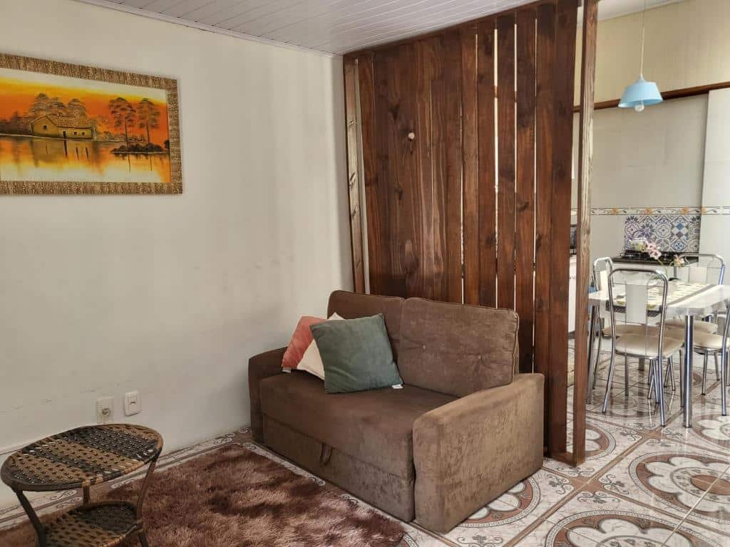Sala e cozinha no Residência Cardoso. Há um sofá de dois lugares, e atrás dele, um painel de madeira separando a área da sala para a cozinha. Na cozinha, vemos parcialmente uma mesa com quatro cadeiras.