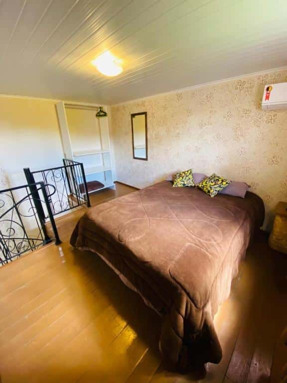 Quarto do RESIDENCIAL BORGES CANELA. Há uma cama de casal no centro, e à sua esquerda há uma estante com cabides e um espelho. No topo da foto à direita vemos um ar-condicionado. Uma escada que dá acesso direto ao quarto está à esquerda, no pé da cama. Representa o post sobre airbnb em Canela.
