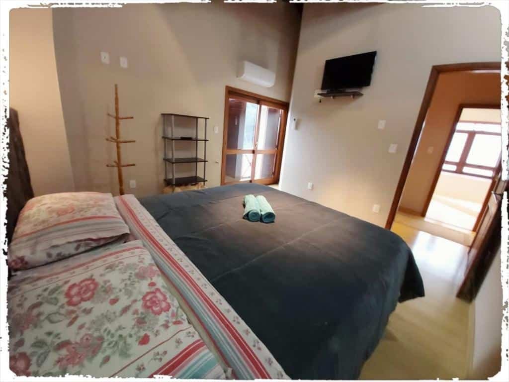 Quarto no Residencial João Pessoa. A cama está no meio da foto, e vemos ela do ângulo como quem está deitado. Na sua frente há uma TV fixa na parede e uma porta para o corredor. No fundo à esquerda há uma porta para a varanda e um ar-condicionado acima. Representa o post sobre airbnb em Canela.