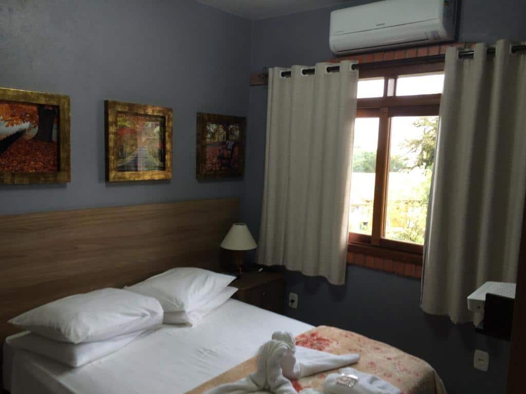 Quarto no Residencial Quatro Estações. Há uma cama box de casal no canto inferior esquerdo, e uma cabeceira de madeira atrás com abajur. No seu lado direito há uma janela e ar-condicionado. Representa o post sobre airbnb em Canela.