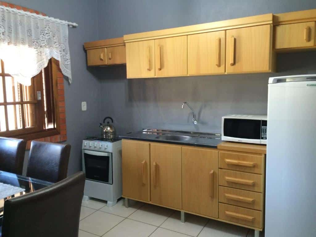 Cozinha do Residencial Quatro Estações. No canto inferior esquerdo vemos parcialmente uma mesa com cadeiras de couro. Na frente dela, há vários armários, uma pia, fogão, micro-ondas e geladeira.