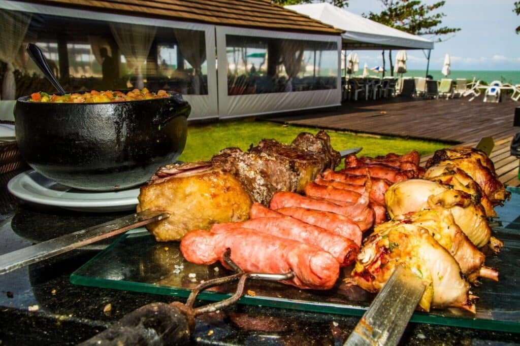 Espetos de carne grelhada e salsichas são expostos em uma mesa ao ar livre no Resort Arcobaleno All Inclusive, perto de uma área de jantar coberta com vista para o mar.