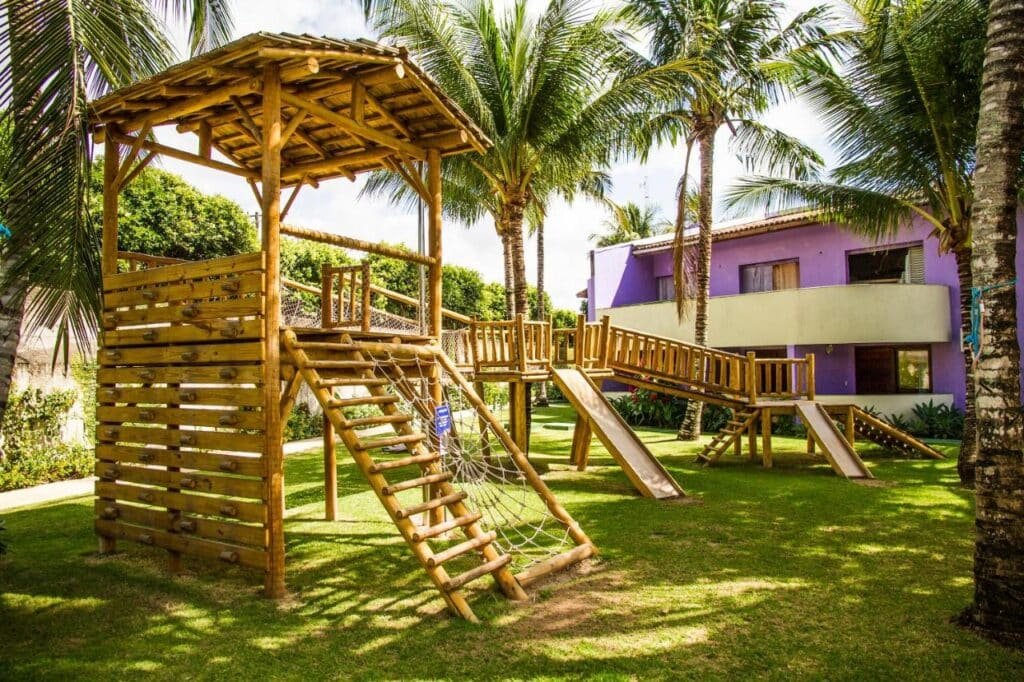 Um playground de madeira com estruturas de escalada e escorregadores fica em uma área gramada, cercado por palmeiras e um prédio roxo no Resort Arcobaleno All Inclusive.