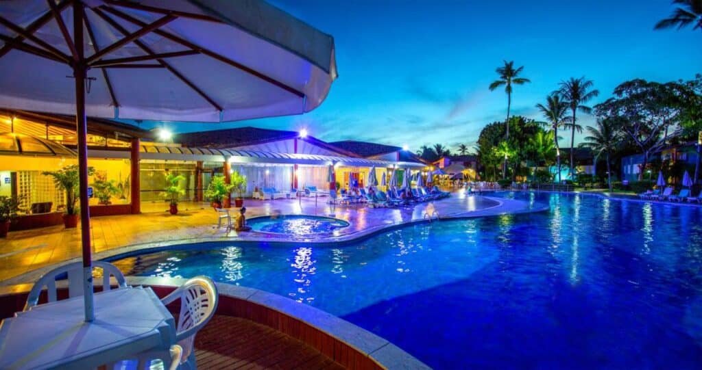 Uma área de piscina do Resort Arcobaleno All Inclusive ao anoitecer, com uma grande piscina, espreguiçadeiras, árvores e um edifício com janelas iluminadas sob um céu limpo.