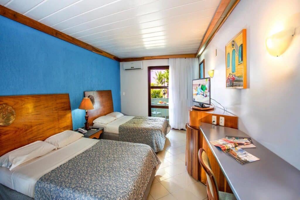 Quarto do Resort Arcobaleno All Inclusive com duas camas de casal, uma parede com detalhes em azul, uma mesa de madeira com TV e uma varanda com vista para palmeiras do lado de fora.