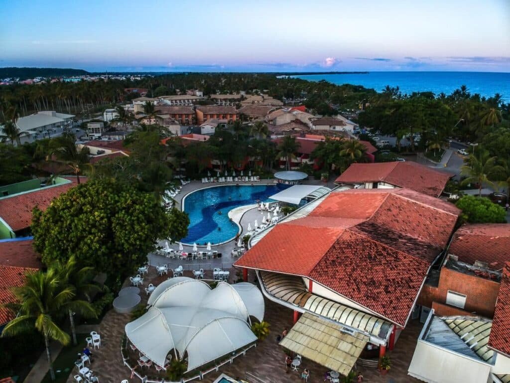 Vista aérea do Resort Arcobaleno All Inclusive com edifícios com telhados vermelhos, uma grande piscina e palmeiras perto do oceano ao pôr do sol.