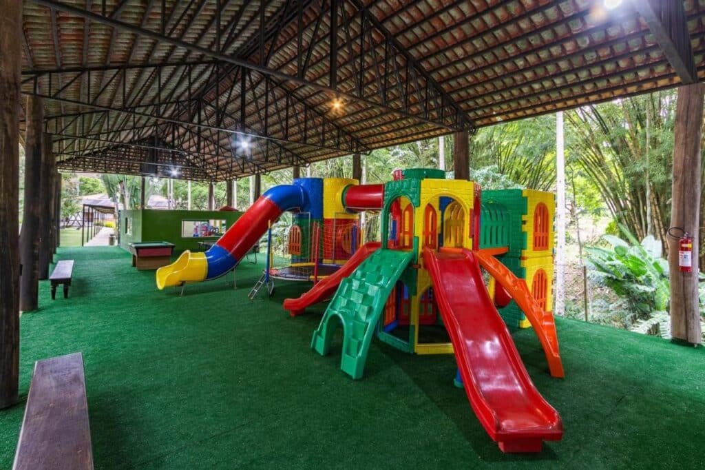 Parque infantil coberto com escorregadores coloridos e estruturas de escalada sob um teto de madeira, cercado por piso de grama artificial no Hotel Fazenda 3 Pinheiros.