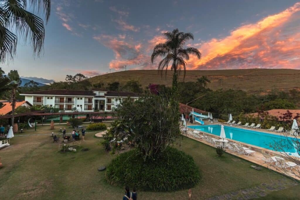 Hotel Fazenda 3 Pinheiros, um dos hotéis fazenda no Rio de Janeiro, com piscina, espreguiçadeiras e jardim cercado por colinas ao pôr do sol. Os hóspedes ficam espalhados pela área gramada.