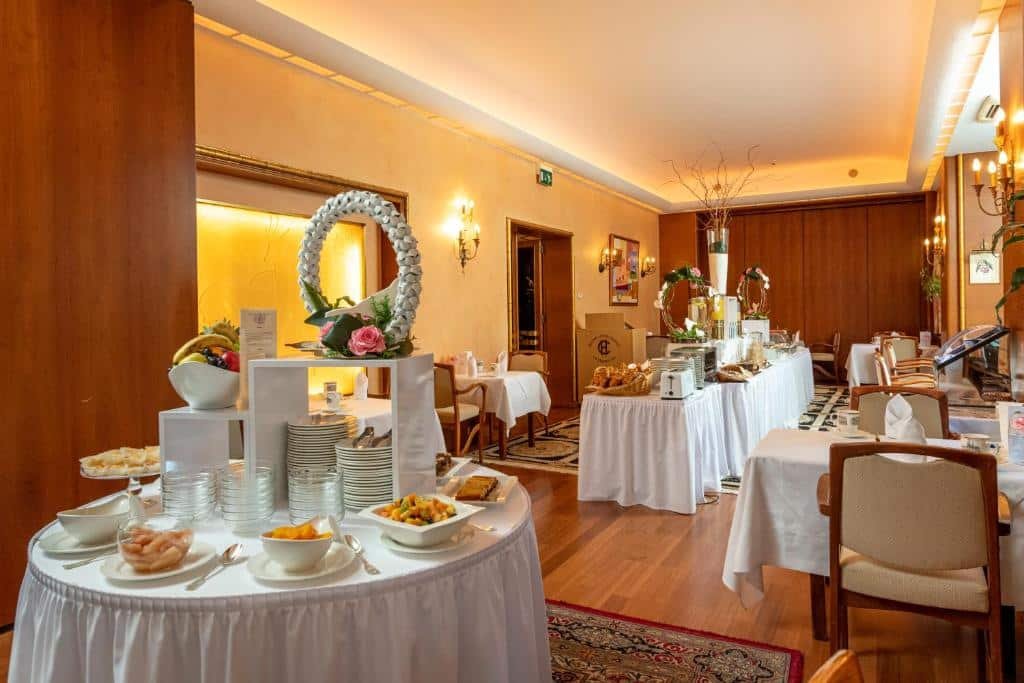 Restaurante do Grand Hotel Cravat servindo um buffet. Em primeiro plano, temos uma mesa redonda com lençóis brancos, que dispõe tigelas de vidro e porcelana com alimentos dentro, e no centro, estão os pratos. Caminhando mais ao fundo, há outra mesa, dessa vez retangular, que possui mais variedades de alimentos. Encostadas nas paredes, estão as mesas com cadeiras. O ambiente é decorado com tons terrosos, alguns painéis de madeira e candedalabros.