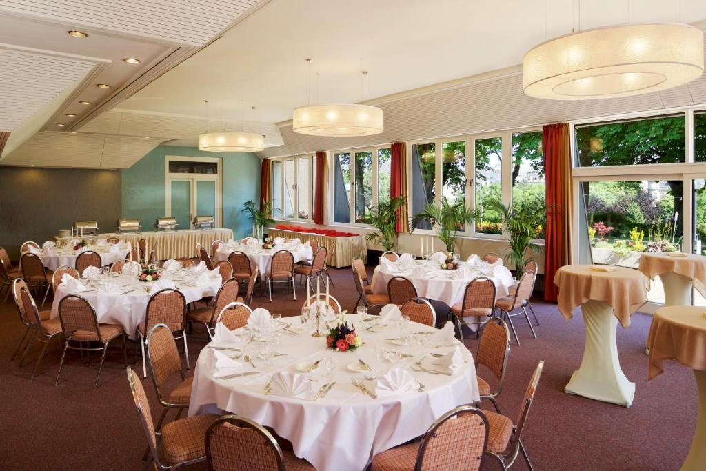 Restaurante do hotel Parc Plaza. O ambiente é clássico e elegante. As mesas são redondas e possuem nove cadeiras. Estão arranjadas com toalhas brancas e talheres, e flores no centro de cada uma. O teto recebe lustres grandes redondos. Através da janela, vemos várias árvores. O piso está coberto por um carpete roxo.