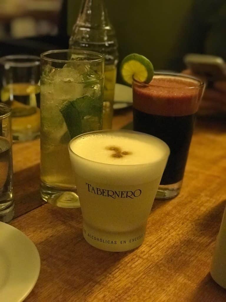 três copos de bebidas do Restaurante Inka Grill. Um copo de pisco sour, um copo de chicha morada e uma garrafa de vidro de inka cola