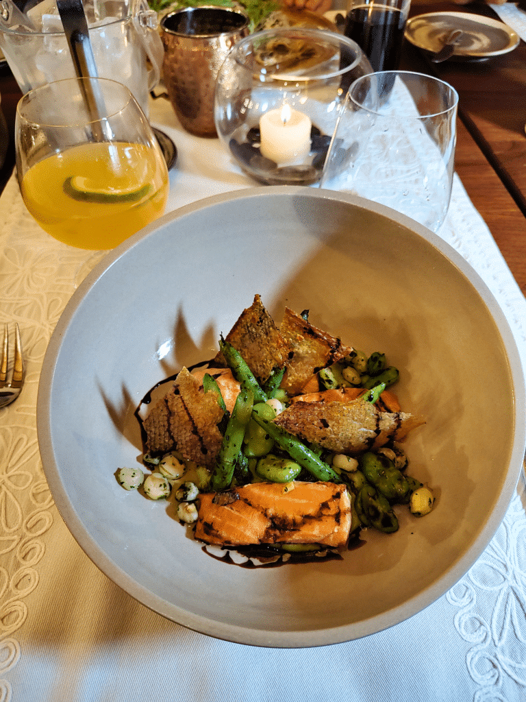 prato fundo arredondado e cinca do restaurante el huerto, com salmão grelhado, milho e quinoa, e aspargos. Há também dois copos de vidro, um de cada lado do copo, no lado esquerdo há um líquido alaranjado com uma rodela de laranja dentro. 