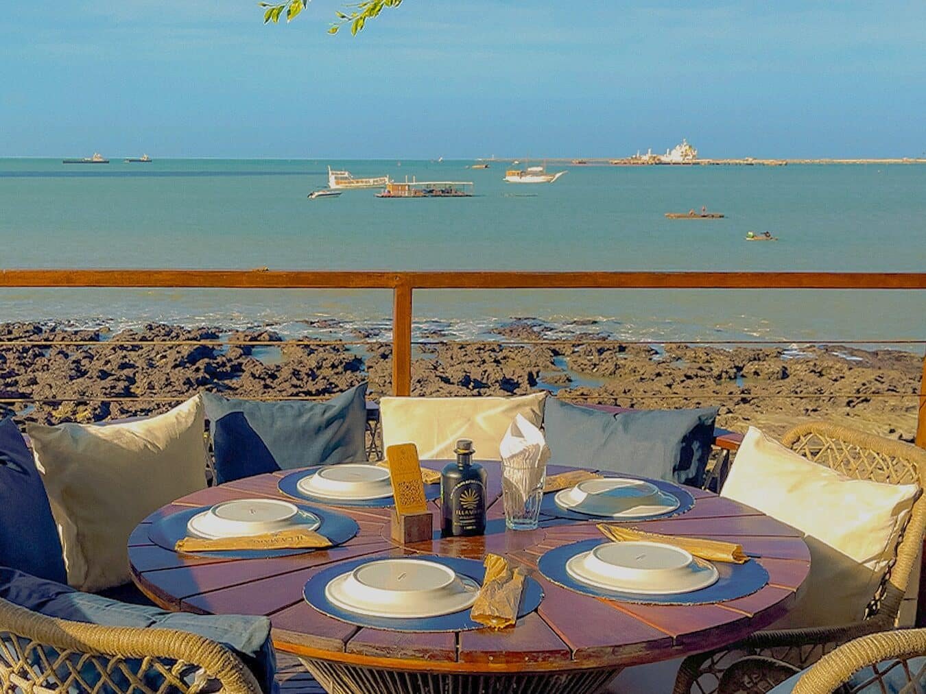 Foto de uma mesa do restaurante Illa Mare em uma varanda beira-mar. A vista é para a praia, que contém  algumas embarcações passando. O céu está azul e sem nuvens. A mesa está posta com pratos e talheres e há assentos com almofadas.