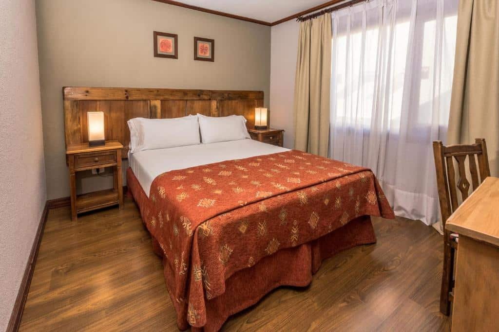 Quarto do Rincón del Calafate. Uma cama de casal, de cada lado uma mesinha de apoio e um abajur. De frente uma mesa com uma cadeira e do lado direito a janela com cortina. Foto para ilustrar post sobre hotéis em El Calafate.