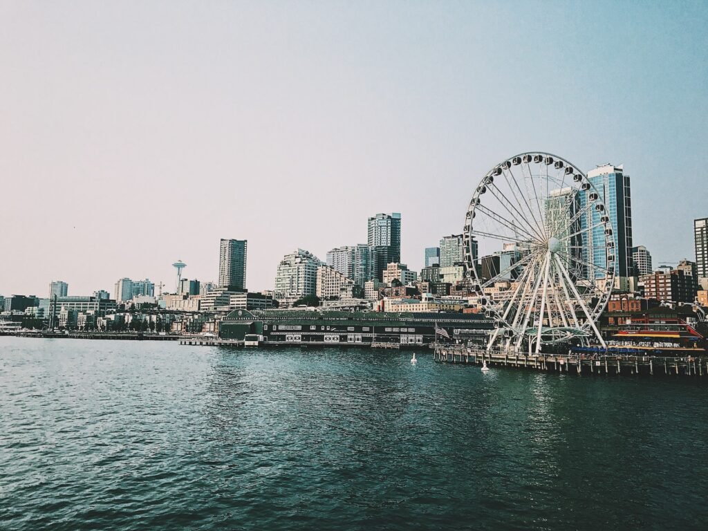 fotografia da costa de Seattle mostrando a roda gigante no píer e diversos prédios modernos ao fundo. Na parte debaixo da imagem há o mar escuro e na parte de cima, um céu claro