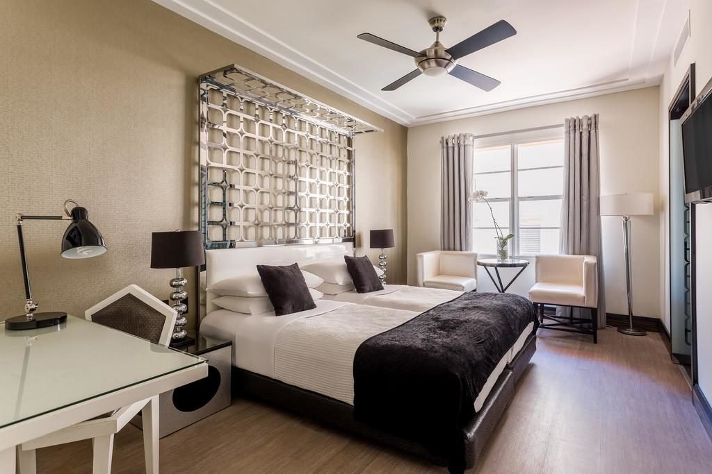 A imagem mostra um quarto de hotel elegante e bem decorado no Waldorf Towers. Há duas camas de solteiro com roupa de cama branca e colchas pretas, decoradas com almofadas pretas. Acima das camas, uma cabeceira decorativa em metal com design geométrico adiciona um toque sofisticado. À esquerda, há uma mesa de trabalho branca com uma luminária moderna. Perto da janela, que tem cortinas cinza, há duas poltronas brancas com uma mesa de vidro entre elas, adornada com uma orquídea. No teto, um ventilador de teto moderno completa o ambiente aconchegante e funcional.