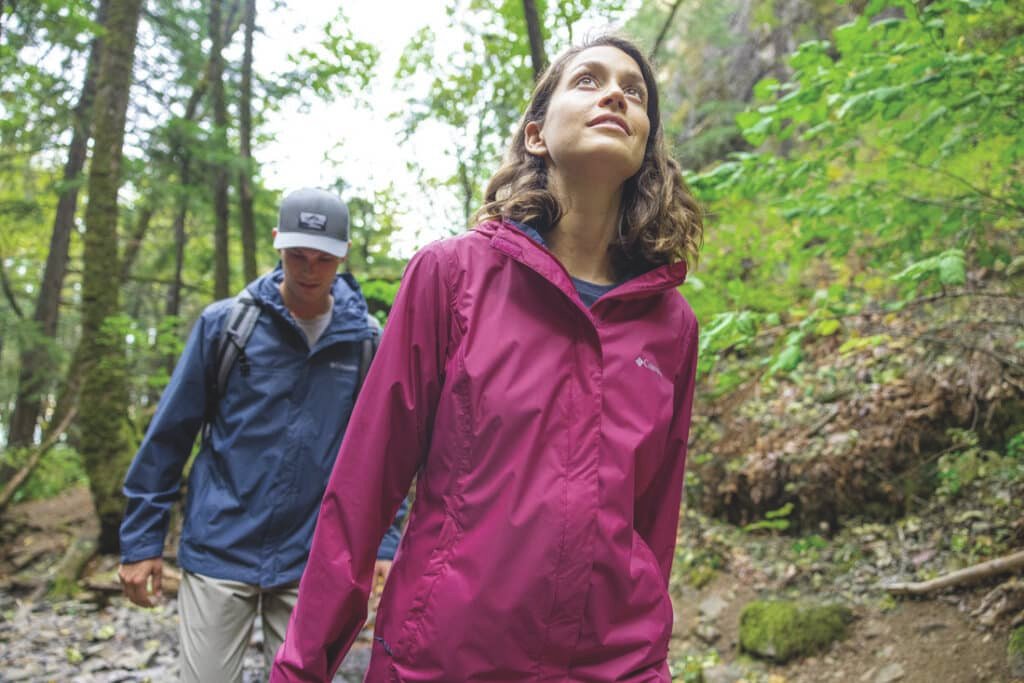 Duas pessoas caminhando com roupa de trilha em uma floresta. A mulher na frente usa uma jaqueta vermelha, o homem atrás usa uma jaqueta azul e boné.