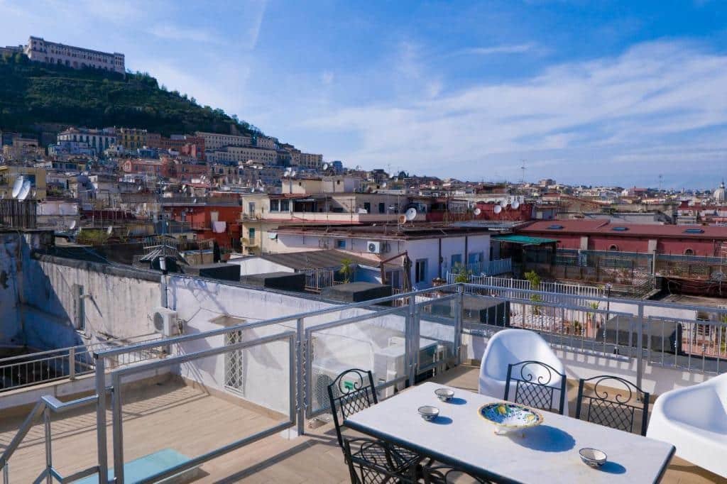 Terraço do Royal Boutique Napoli com uma mesa com três cadeiras, poltronas brancas e uma vista durante o dia para as construções da cidade, ilustrando post Hotéis em Nápoles.