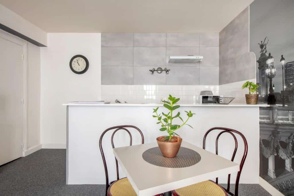 Cozinha de estúdio no Royal Hotel Versailles. Uma mesinha com duas cadeiras e um vaso de plantas está no centro do cômodo, e ao fundo um balcão separa a sala da cozinha, onde pe possível ver um micro-ondas e um exaustor. Ao lado esquerdo há um relógio.