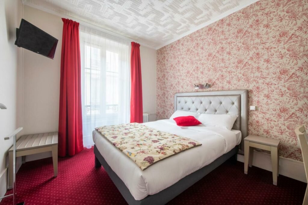 Quarto do Royal Hotel Versailles, um dos hotéis perto do Palácio de Versalhes. Uma cama de casal encostada na parede direita tem mesinhas de cabeceira dos dois lados e encara uma TV na parede oposta. Ao lado da cama há uma janela com cortinas.