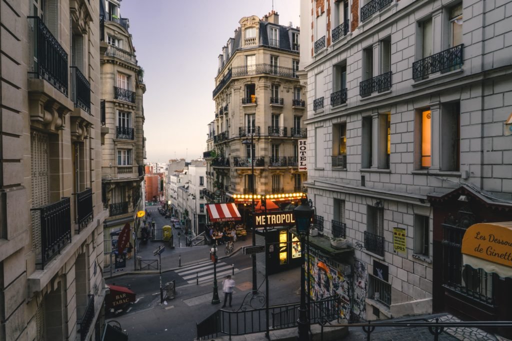 Uma rua de Montmartre, em Paris, com edifícios clássicos adornados com varandas de ferro forjado. Ao centro, um letreiro iluminado do "Métropol" e uma fachada de um café com toldo listrado de vermelho e branco, trazendo um toque vintage. As janelas iluminadas e as escadarias de ferro adicionam charme ao cenário, que parece estar ao entardecer.