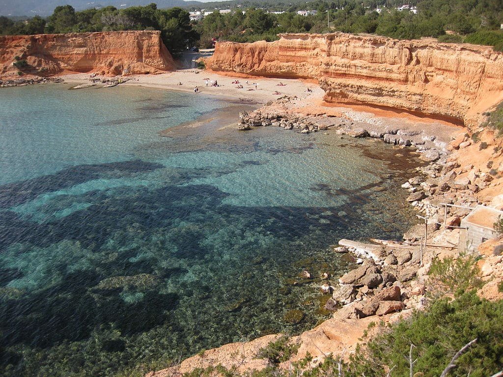 Litoral rochoso com água azul-turquesa clara e uma pequena praia de areia. Pessoas tomando sol na praia. Arbustos em primeiro plano.