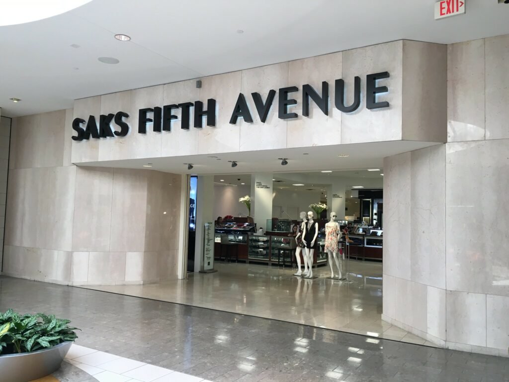 Entrada e faixada da loja Saks Fifth Avenue dentro do shopping Dadeland Mall. Ela está aberta e tem alguns manequins na entrada. Mais ao fundo estão alguns balcões com vitrines.