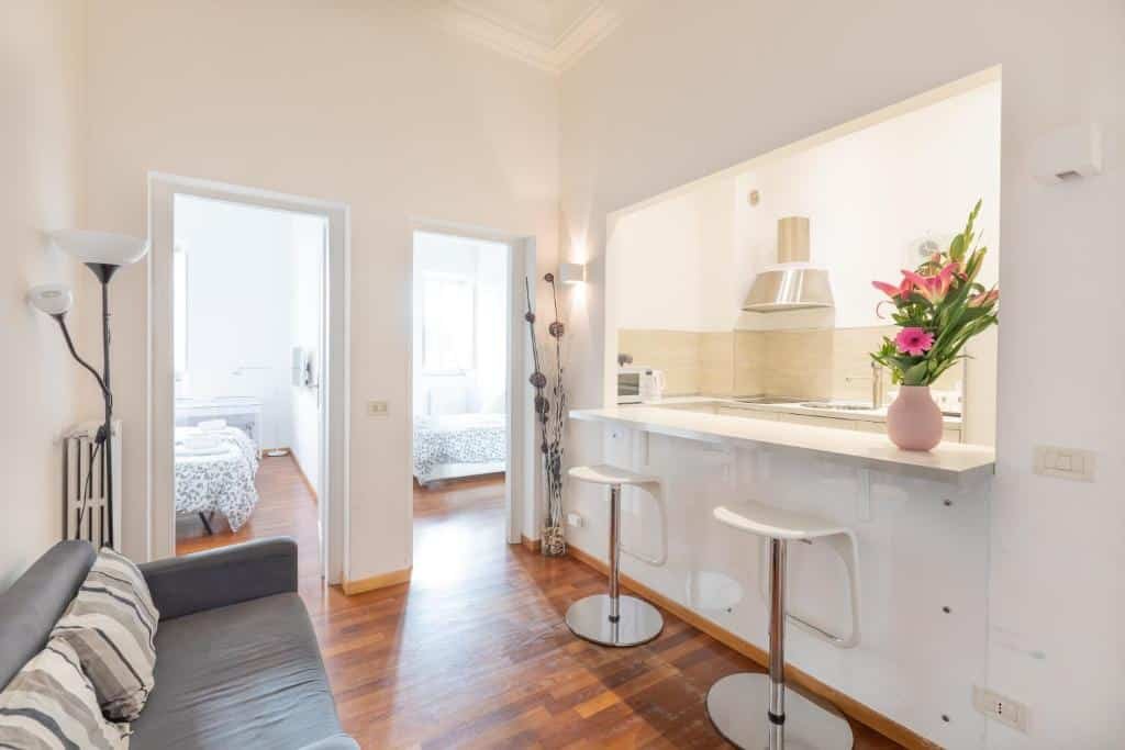 sala do Stay In Rome com sofá e cozinha americana à frente, com duas cadeiras altas e equipamentos de cozinha, há duas portas que dão para os dois quartos do apartamento