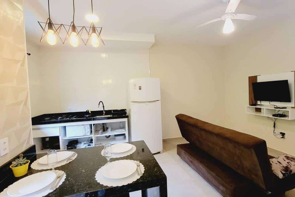 Sala de estar do Apê na praia da Lagoinha a 400m da praia com mesa de mármore a frente do lado esquerdo da imagem e ao fundo uma pia com geladeira, do lado direito um sofá e em frente ao sofá um painel com TV. Representa airbnb na praia da Lagoinha.