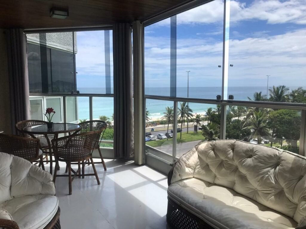 Varanda do Vistão do mar – posto 5 Barra da Tijuca com sofá do lado direito e ao fundo uma mesa redonda com quatro cadeiras com vista para a praia.