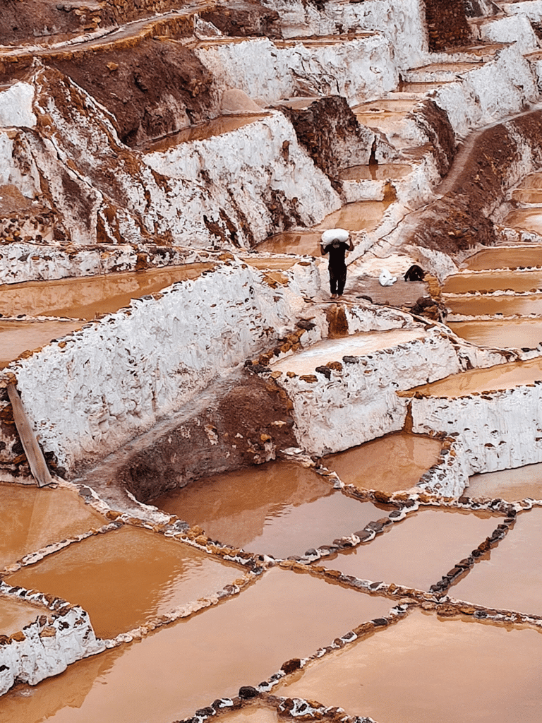 trabalhado carregando uma saco de sal nas costas no lado direito da imagem. Ele está caminhando entre as piscinas de sal da Salineras de Maras, no Vale Sagrado dos Incas.