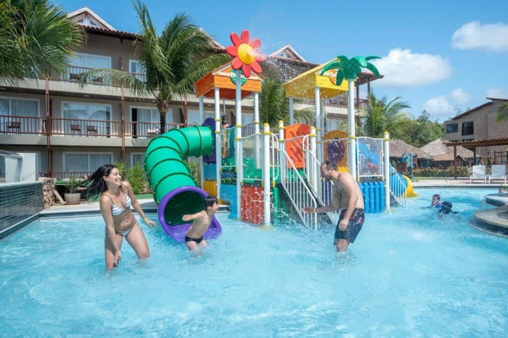 Pessoas aproveitando um playground aquático com toboáguas e estruturas coloridas na área da piscina do Salinas Maceio All Inclusive Resort sob um céu azul claro.