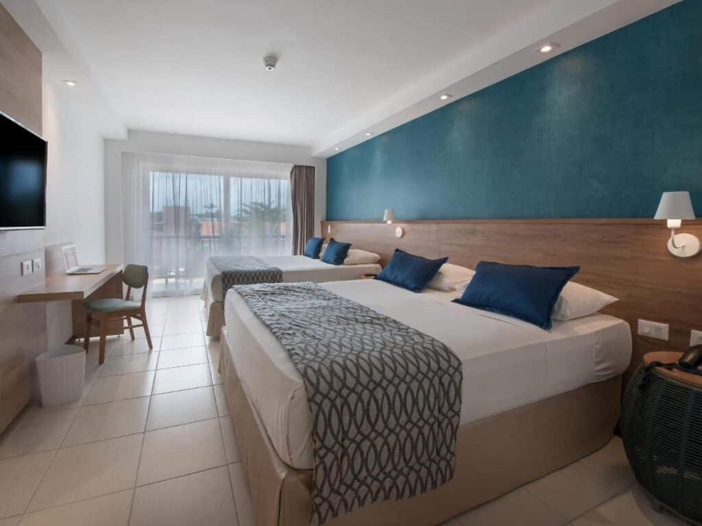 Quarto do Salinas Maceio All Inclusive Resort com duas camas de casal, parede com detalhes em azul, TV montada na parede, escrivaninha com cadeira e uma grande janela com cortinas.