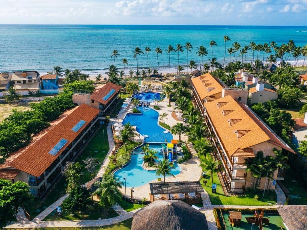 Vista aérea do Salinas Maceio All Inclusive Resort com dois edifícios e uma grande piscina perto de uma praia arenosa cercada por palmeiras.