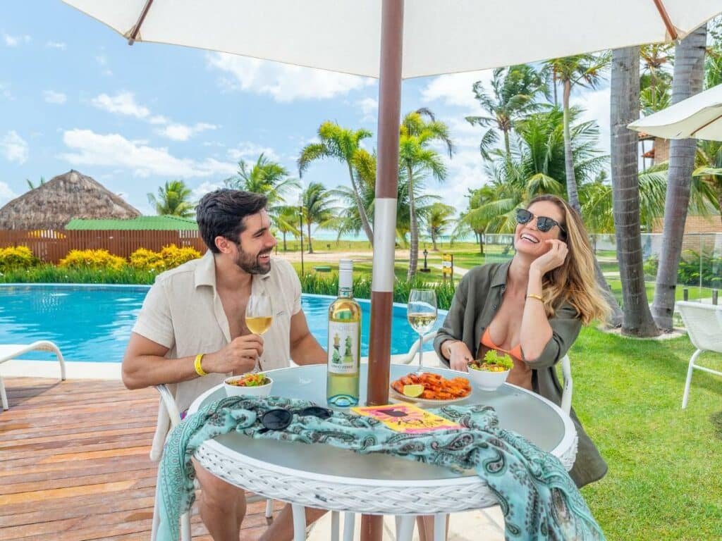 A imagem mostra um casal sentado em uma mesa ao ar livre, ao lado de uma piscina no Salinas Maragogi All Inclusive Resort, um dos melhores resorts all inclusive no Brasil. Ambos estão sorrindo e aproveitando uma refeição leve, com taças de vinho branco. O homem veste uma camisa bege e a mulher usa óculos escuros, um biquíni laranja e uma camisa verde aberta. Sobre a mesa, há um prato de camarões e uma salada, junto com uma garrafa de vinho. Ao fundo, há palmeiras, um céu azul com poucas nuvens, e uma cabana de palha, realçando a atmosfera relaxante e ensolarada do local.