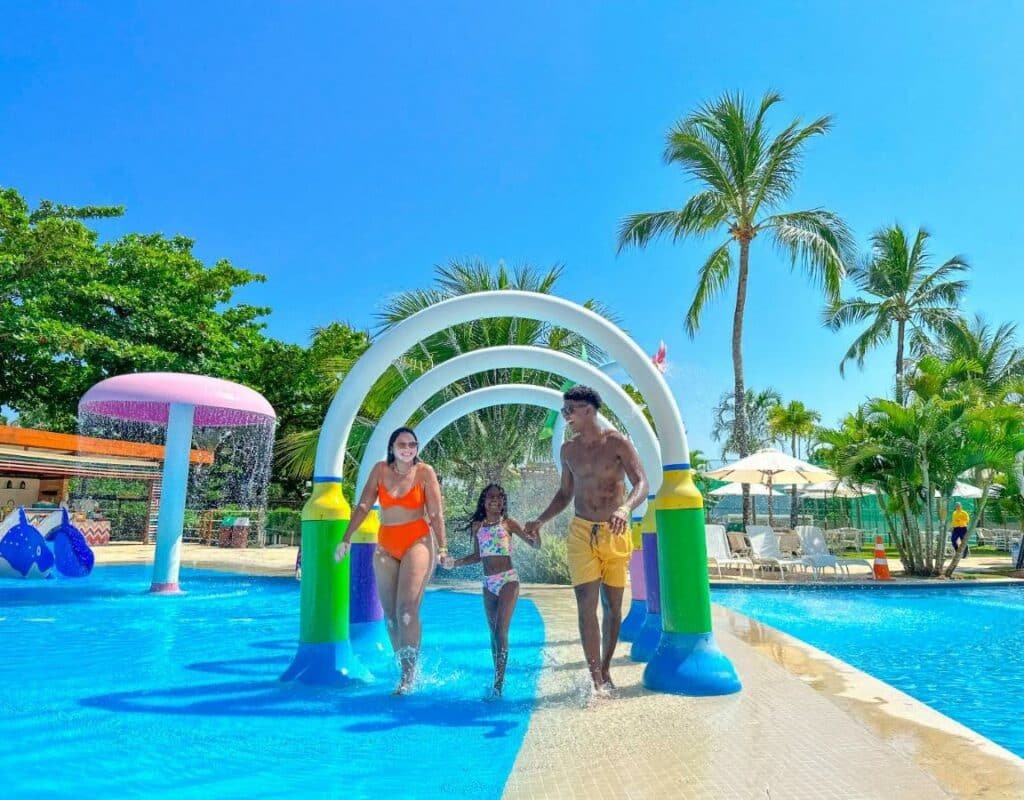 A imagem mostra uma família se divertindo em um parque aquático ao ar livre no Salinas Maragogi All Inclusive Resort, um dos melhores resorts all inclusive no Brasil. A mãe, vestindo um maiô laranja, o pai, usando um short de banho amarelo, e a filha, com um biquíni colorido, estão correndo juntos sob arcos coloridos que jorram água, criando um efeito de túnel de água. Ao fundo, há mais estruturas de brinquedos aquáticos, como um grande cogumelo rosa que também esguicha água.