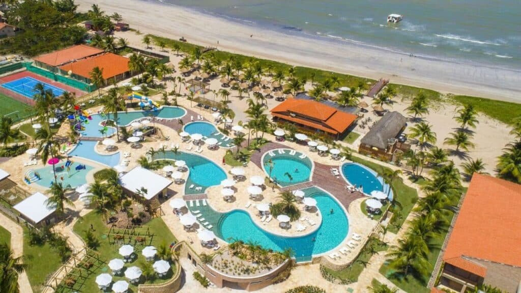 A imagem mostra uma vista aérea do Salinas Maragogi All Inclusive Resort com várias piscinas de formatos diferentes rodeadas por espreguiçadeiras e guarda-sóis brancos. Há uma área de parque aquático com escorregadores coloridos e uma piscina infantil. O resort também inclui quadras de tênis e futebol, visíveis à esquerda da imagem. Ao fundo, uma extensa faixa de praia com areia clara se estende até o mar, onde um pequeno barco navega próximo à costa. O cenário é repleto de palmeiras, proporcionando uma atmosfera tropical e convidativa para relaxamento e lazer.