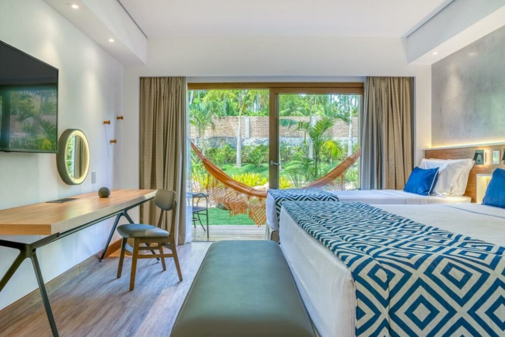 A imagem mostra um quarto do Salinas Maragogi All Inclusive Resort. Há duas camas com colchas brancas e almofadas azuis, uma mesa de trabalho com uma cadeira e um espelho redondo iluminado. Através de uma grande porta de vidro, pode-se ver uma varanda com uma rede laranja, cercada por um jardim verde com palmeiras e vegetação tropical. A decoração é clean e sofisticada, com cores claras e detalhes em madeira.